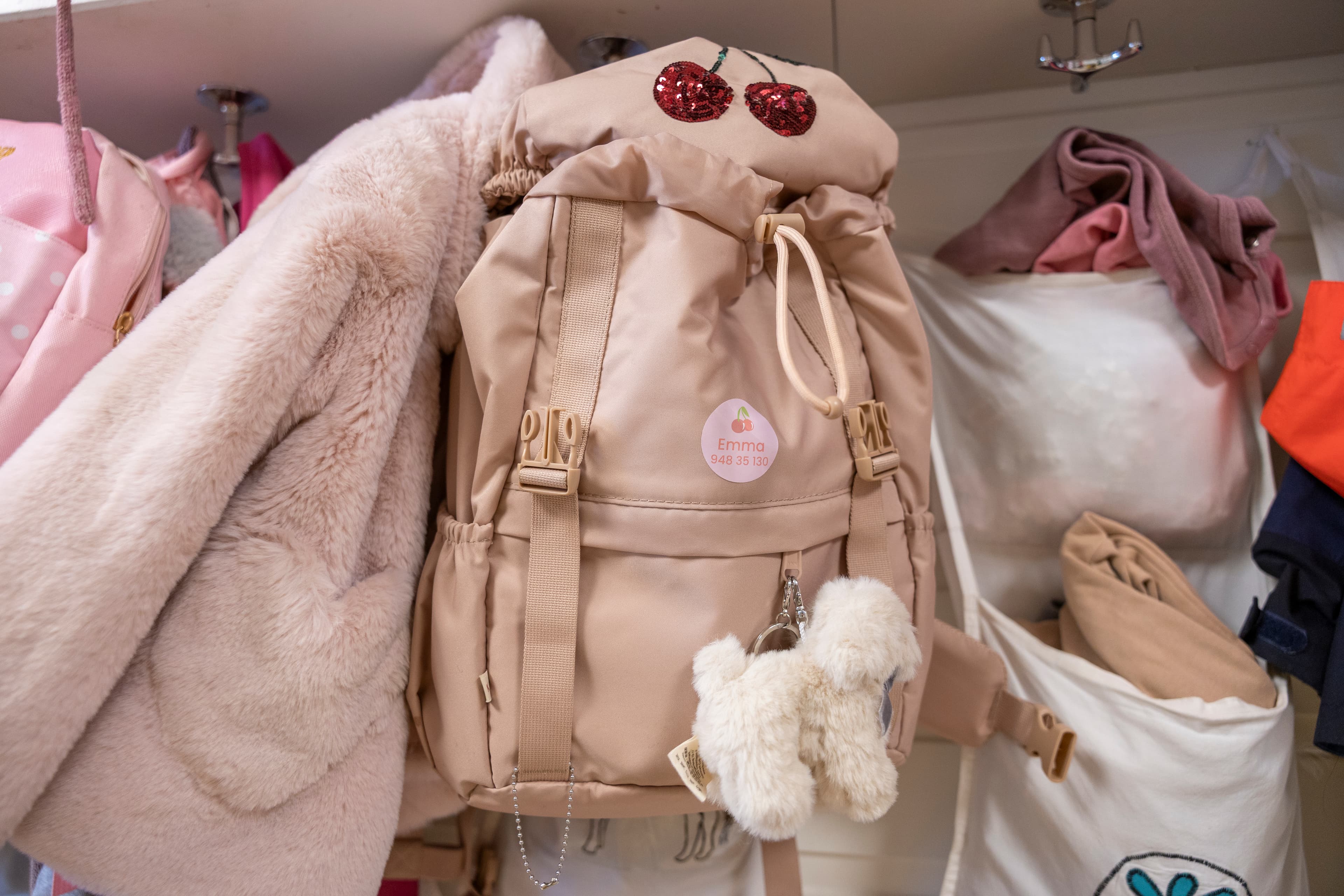 Beige sekk merket med en rosa navnelapp, hengt opp i en barnehage- eller skolegarderobe blant andre yttertøy og bager.