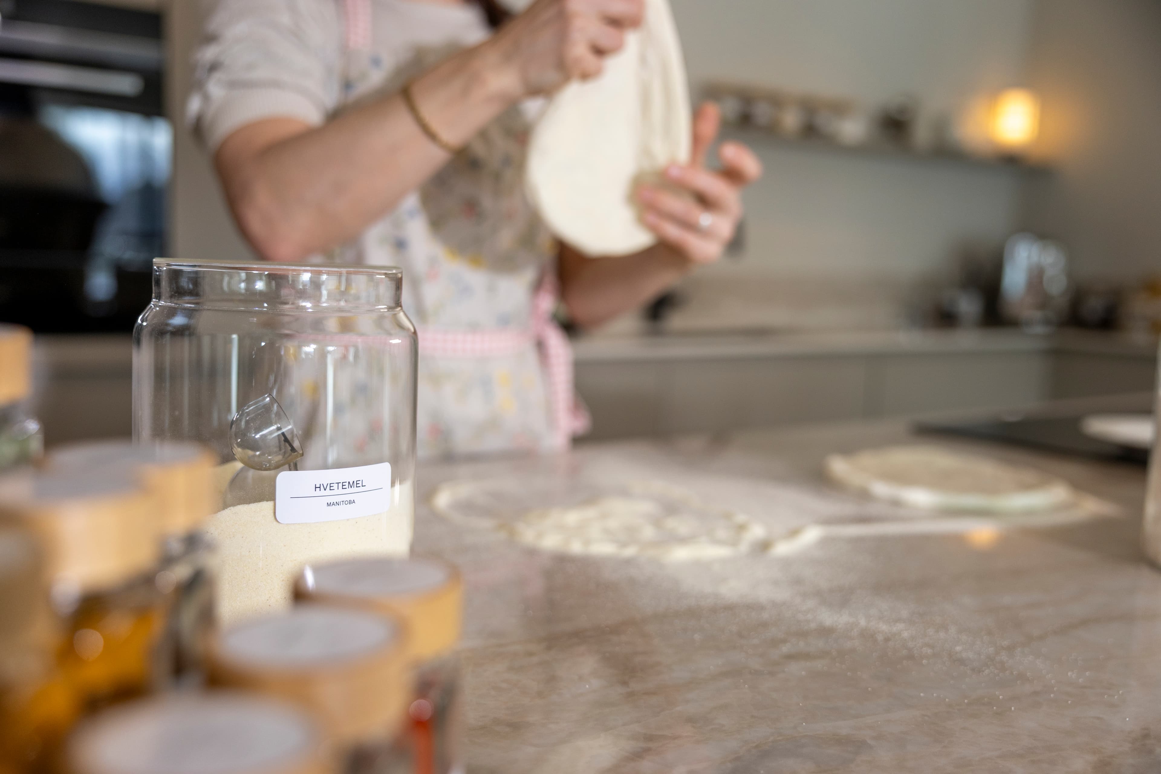Kvinne baker brød på et kjøkken. På benken står glasskrukker med mel merket med etiketter, og et nybakt brød ligger fremme.