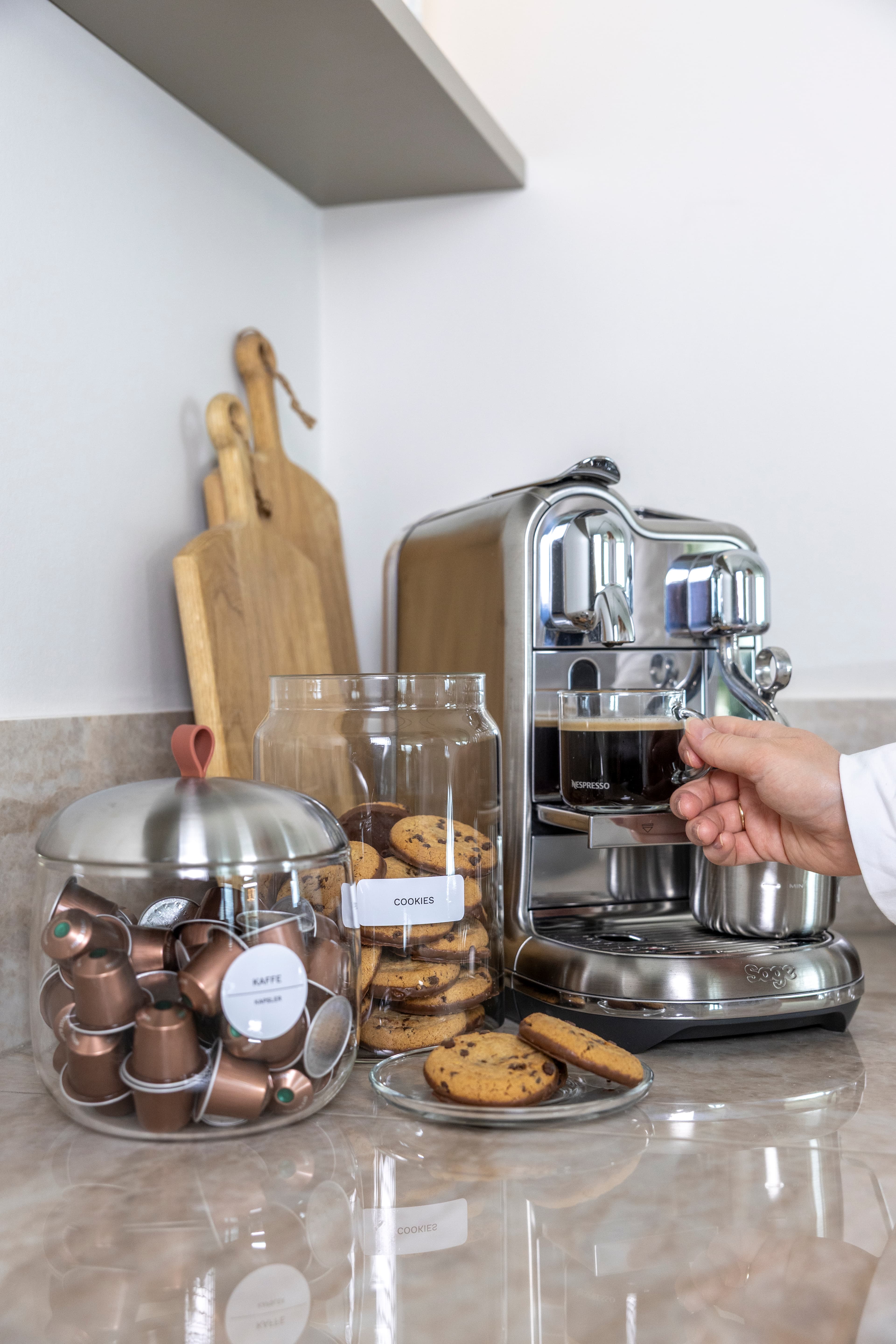 Glasskrukke med kaffekapsler merket «Kaffe» og en krukke med småkaker merket «Cookies» ved siden av en kaffemaskin.
