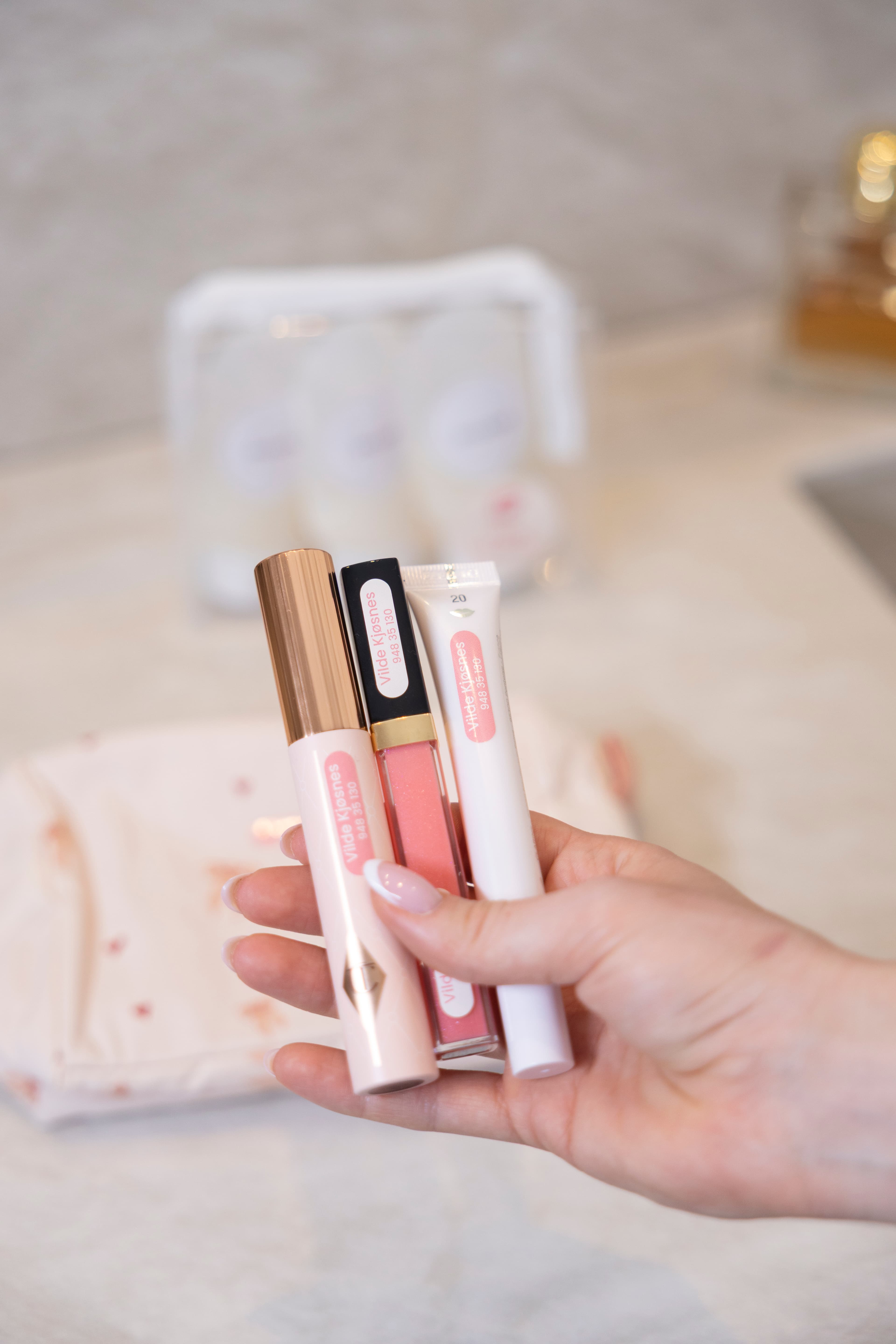 Hånd holder tre leppeprodukter – en leppestift, en lipgloss og en leppebalsam – alle merket med små navnelapper.