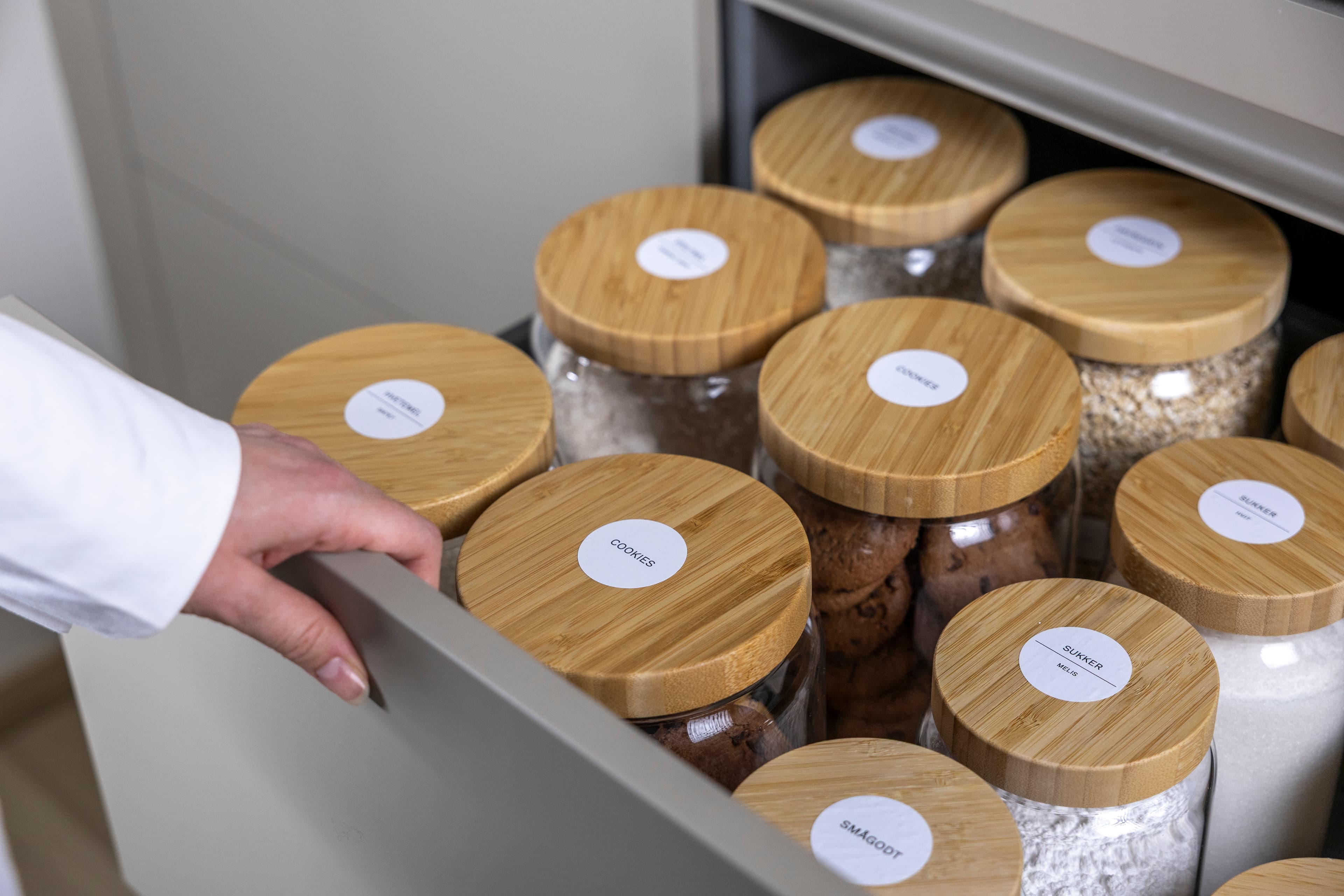Hånd åpner en kjøkkenskuff med oppbevaringskrukker merket «Cookies», «Sukker», «Hvetemel» og «Smågodt».